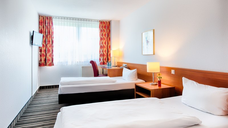 Helles Zweibettzimmer mit zwei Einzelbetten, Nachttisch mit Lampe, Schreibtisch, Fernseher und roten Vorhängen., © ACHAT Hotels Deutschland