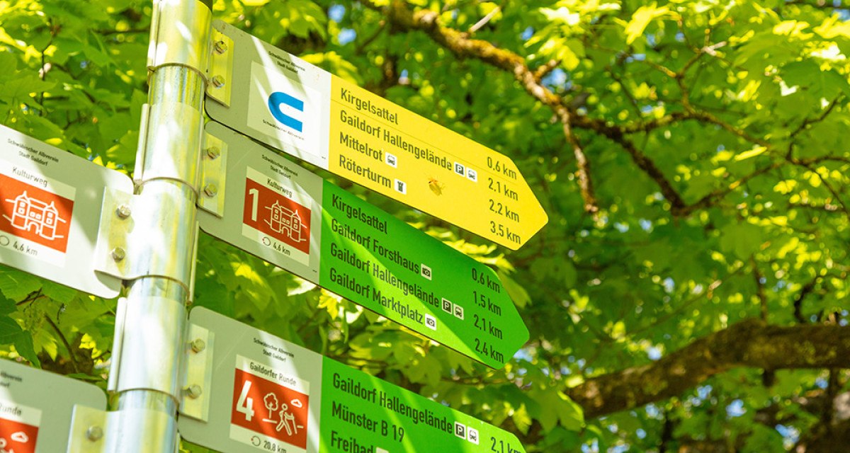 Wegweiser für Rundwanderwege in Gaildorf mit Entfernungsangaben, umgeben von grünem Laub. Wegweiser für Rundwanderwege in Gaildorf mit Entfernungsangaben, umgeben von grünem Laub.