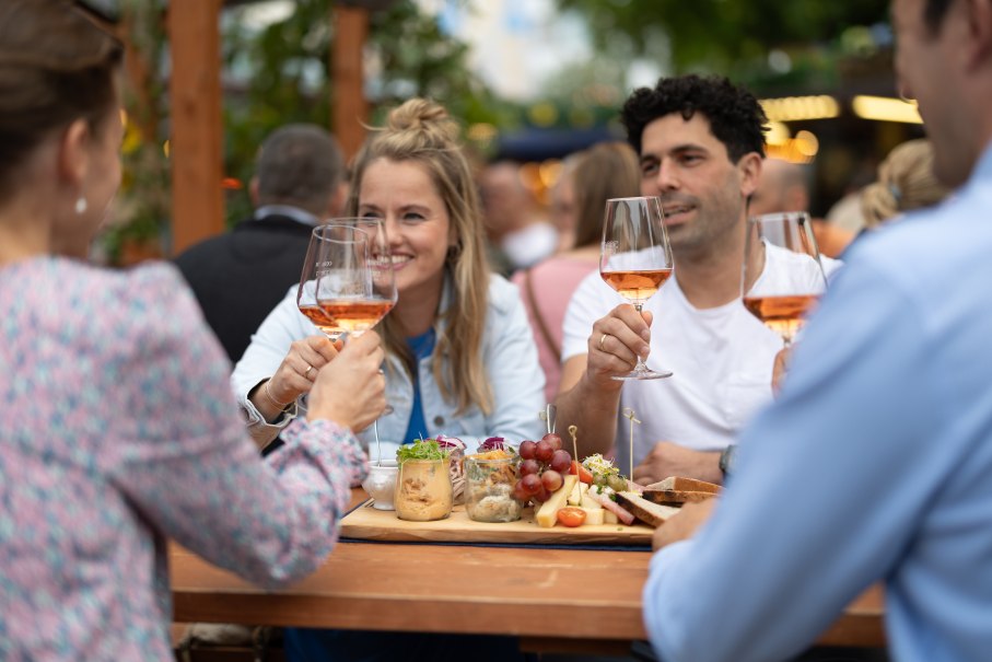 Vier Personen stoßen mit Weingläsern an einem Tisch im Freien an, umgeben von Essen und einer geselligen Atmosphäre., © Stadt Nürtingen, B. Knoblauch