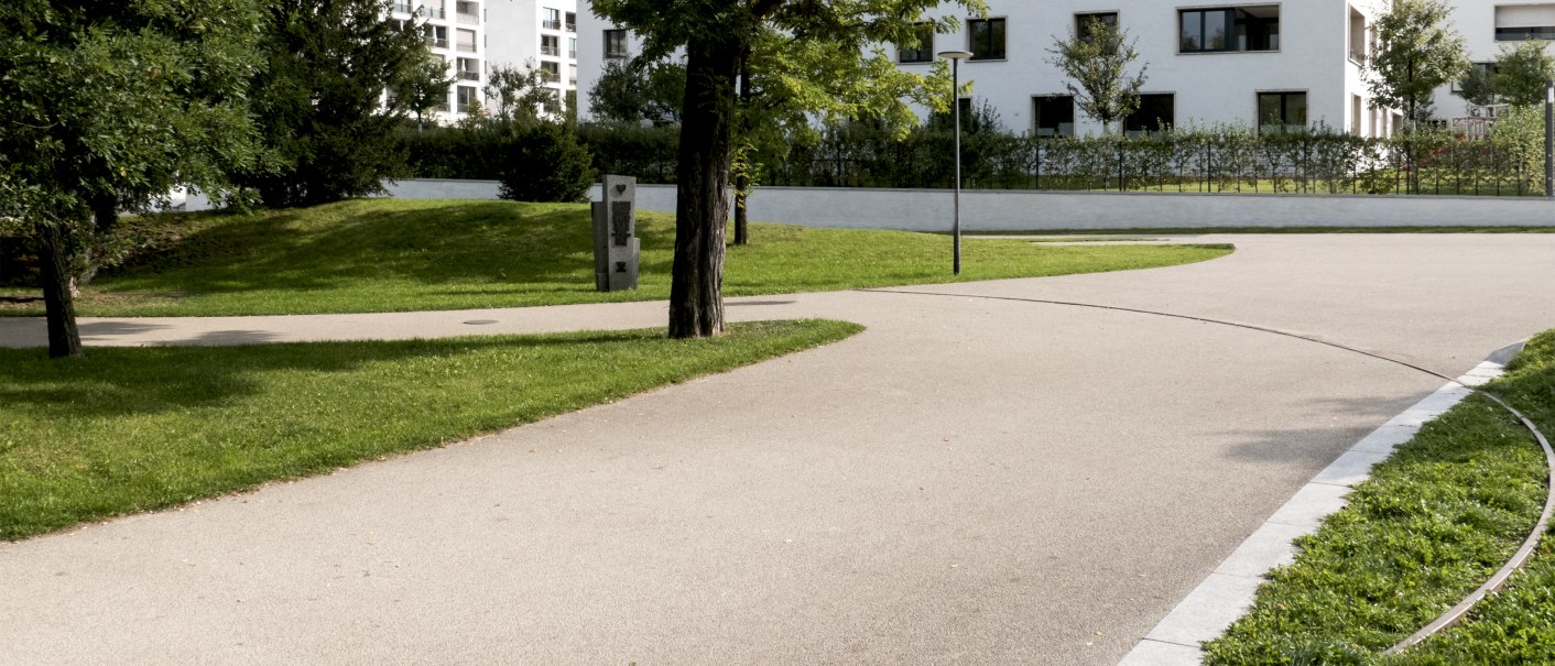 Ein gepflegter Park mit geschwungenen Gehwegen, Bäumen und modernen Wohngebäuden im Hintergrund. Ein Gedenkstein ist sichtbar., © Foto-Kunst Andreas Keller