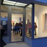 Eine Galerie mit großen Fenstern zeigt bunte Gemälde. Menschen stehen und betrachten die Kunstwerke im Inneren., © EXOgallery Contemporary and New Media Art
