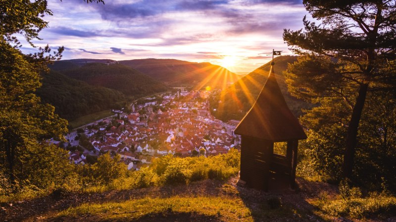 Sonnenuntergang &uuml;ber einem Dorf, gesehen von einem H&uuml;gel mit einer kleinen Holzh&uuml;tte im Vordergrund. Die Sonne strahlt &uuml;ber die Landschaft., &copy; Bad Urach Tourismus