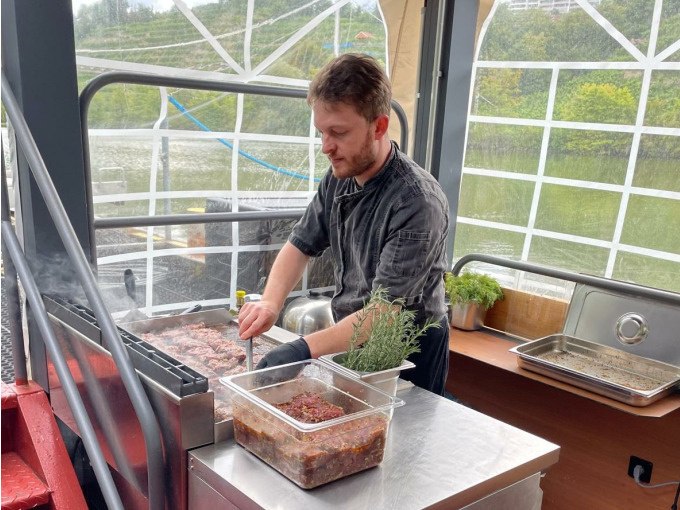 Ein Koch grillt Fleisch auf einem Schiff. Im Hintergrund ist ein Fluss und grüne Landschaft zu sehen. Kräuter und Gewürze stehen bereit., © Cool-Tours StattReisen Ein Koch grillt Fleisch auf einem Schiff. Im Hintergrund ist ein Fluss und grüne Landschaft zu sehen. Kräuter und Gewürze stehen bereit., © Cool-Tours StattReisen