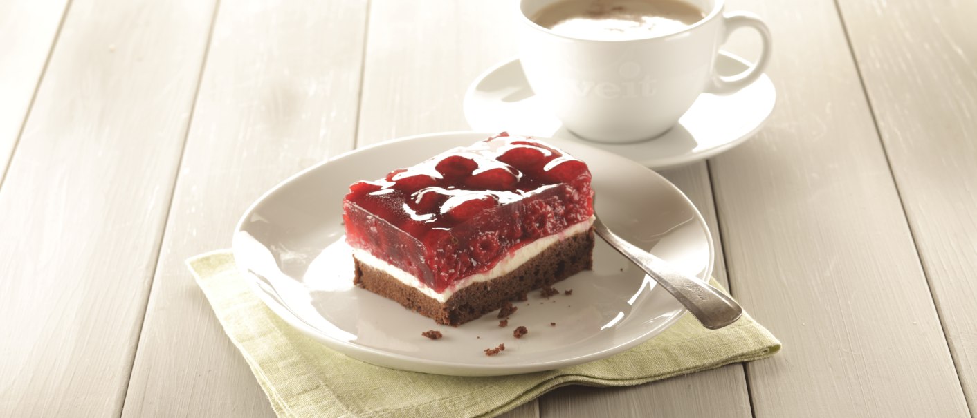 Himbeerschnitte auf einem Teller mit Löffel, daneben eine Tasse Kaffee auf einem Holztisch., © Bäckerei Veit, Victoria Huber