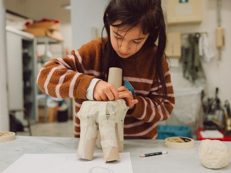 Ein Kind bastelt konzentriert mit Papprollen und Klebeband an einem Tisch in einem Workshop-Raum., © Kunstschule Labyrinth Ein Kind bastelt konzentriert mit Papprollen und Klebeband an einem Tisch in einem Workshop-Raum., © Kunstschule Labyrinth