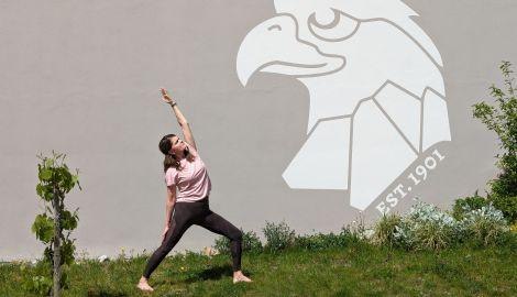 Eine Frau praktiziert Yoga im Freien vor einer Wand mit einem Adler-Logo und der Aufschrift "EST. 1901". Im Vordergrund ist eine kleine Pflanze zu sehen., &copy; Wein & Yoga. Foto: Teamwerk
