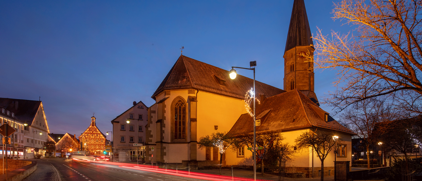 Beleuchtete Kirche bei Dämmerung, umgeben von Bäumen und Gebäuden. Lichtspuren von Autos auf der Straße, festliche Beleuchtung an Laternen., © Stadt Gaildorf