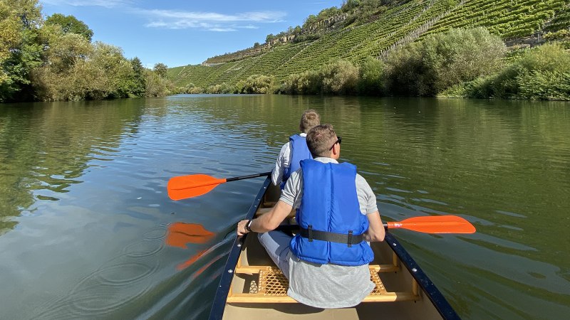Zwei Personen paddeln in einem Kanu auf einem ruhigen Fluss, umgeben von gr&uuml;nen B&auml;umen und Weinbergen unter blauem Himmel., &copy; Matthias Pfl&uuml;ger - Rock-the-River.com