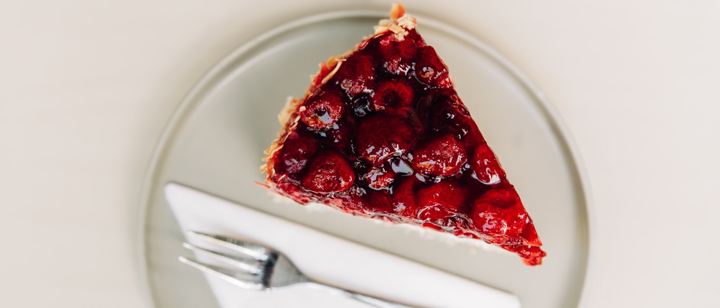Ein Stück Himbeerkuchen auf einem Teller, daneben eine Gabel und eine Serviette. Der Kuchen ist mit glänzenden Himbeeren bedeckt., © L. Prestle/ Documenting Love Ein Stück Himbeerkuchen auf einem Teller, daneben eine Gabel und eine Serviette. Der Kuchen ist mit glänzenden Himbeeren bedeckt., © L. Prestle/ Documenting Love