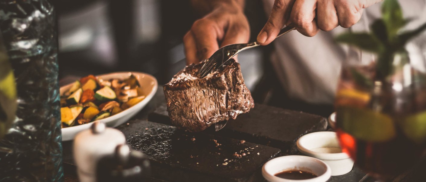 Ein saftiges Steak wird auf einem heißen Stein geschnitten. Im Hintergrund sind Gemüsebeilagen und ein Getränk zu sehen., © Abacco Hotels GmbH Ein saftiges Steak wird auf einem heißen Stein geschnitten. Im Hintergrund sind Gemüsebeilagen und ein Getränk zu sehen., © Abacco Hotels GmbH