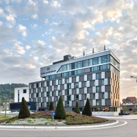 Modernes Hotelgebäude mit Glasfassade, umgeben von einem Kreisverkehr und gepflegter Landschaftsgestaltung, bei Sonnenuntergang., © Hotel am Remspark Modernes Hotelgebäude mit Glasfassade, umgeben von einem Kreisverkehr und gepflegter Landschaftsgestaltung, bei Sonnenuntergang., © Hotel am Remspark