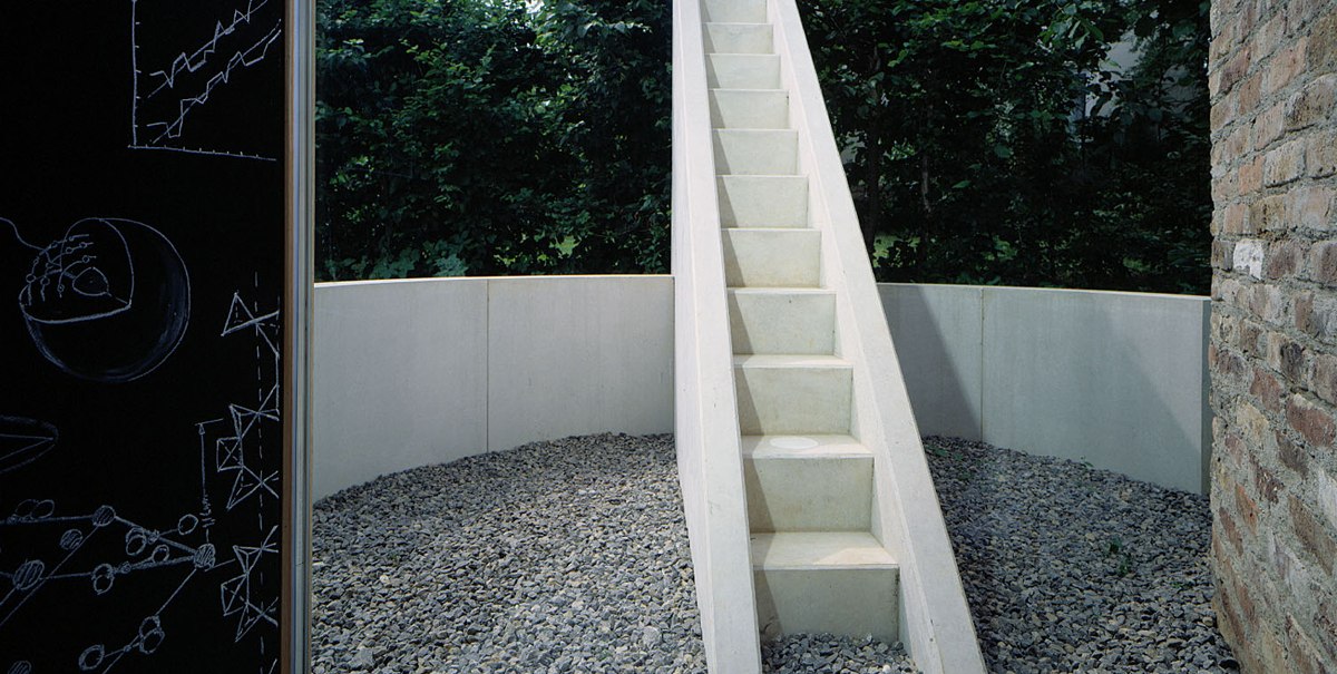 Weiße Treppe in rundem Kiesbereich, umgeben von Bäumen. Links Tafel mit Zeichnungen., © TOMAS