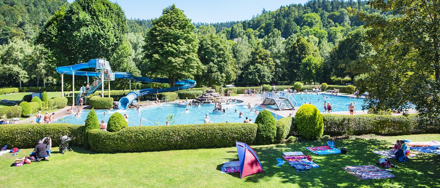Freibad Bad Liebenzell mit blauer Rutsche, Schwimmern im Wasser und grüner Umgebung. Menschen entspannen auf der Wiese unter blauem Himmel., © LOCHER Fotodesing & Manufaktur