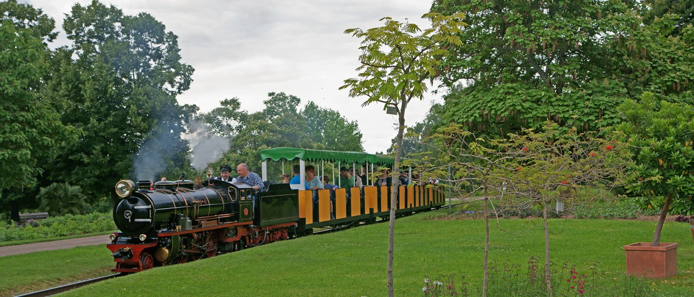 Eine kleine Dampflokomotive zieht bunte Waggons durch einen grünen Park mit Bäumen und Blumen., © Andreas Pucka