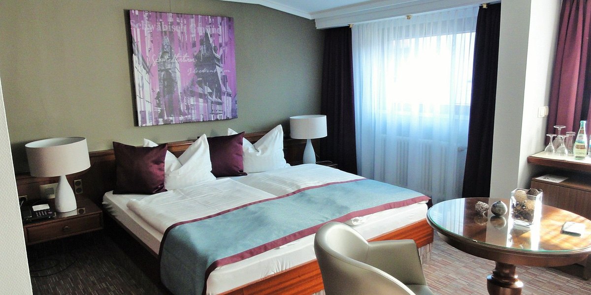 Komfortables Doppelzimmer mit gro&szlig;em Bett, zwei Nachttischen, Tisch und St&uuml;hlen. Helle Vorh&auml;nge und moderne Dekoration an der Wand., &copy; Hotel Pelikan