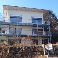 Modernes dreistöckiges Haus mit großen Fenstern und Balkonen, umgeben von Bäumen und blauem Himmel., © Ferienhaus Unteres Remstal