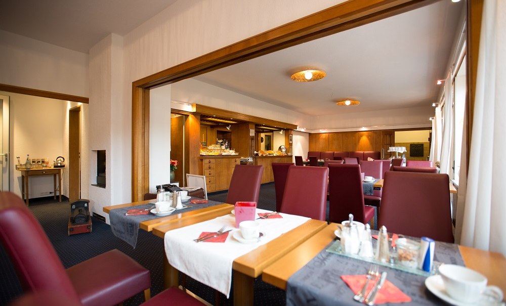 Ein Fr&uuml;hst&uuml;cksraum mit roten St&uuml;hlen, gedeckten Tischen und einem Buffet im Hintergrund. Warme Beleuchtung und Holzelemente schaffen eine einladende Atmosph&auml;re., &copy; Hotel B&uuml;rkle