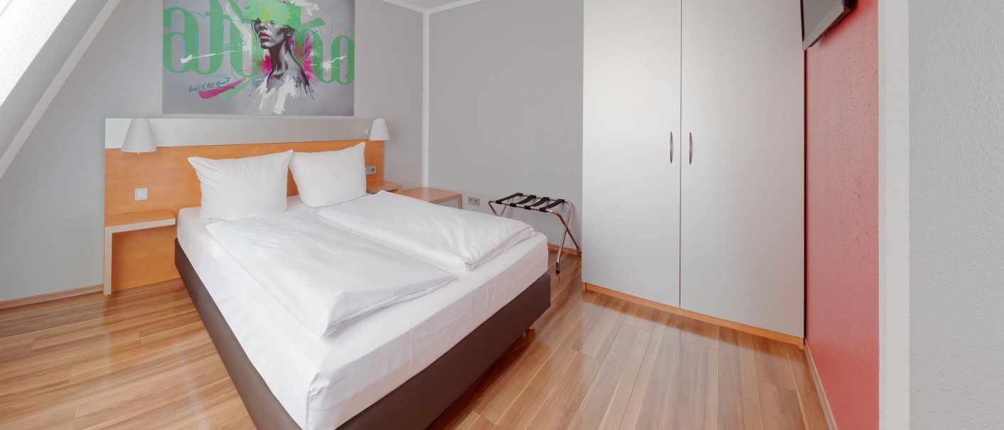 Modernes Hotelzimmer mit Doppelbett, weißem Kleiderschrank, Wandkunst und Flachbildfernseher. Holzboden und rote Akzentwand., © attimo Hotel Stuttgart