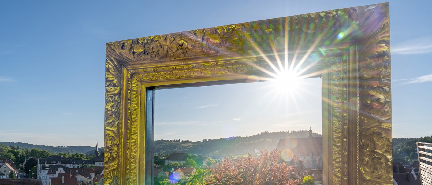 Ein großer, verzierter goldener Bilderrahmen mit Sonnenstrahlen, der einen Blick auf Schwäbisch Gmünd und die umliegende Landschaft bietet., © SMG, Martina Denker