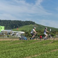 Zwei Radfahrer mit Anhänger fahren durch eine grüne Landschaft in Fellbach. Im Hintergrund sind Hügel und ein modernes Gebäude zu sehen., © Stadt Fellbach