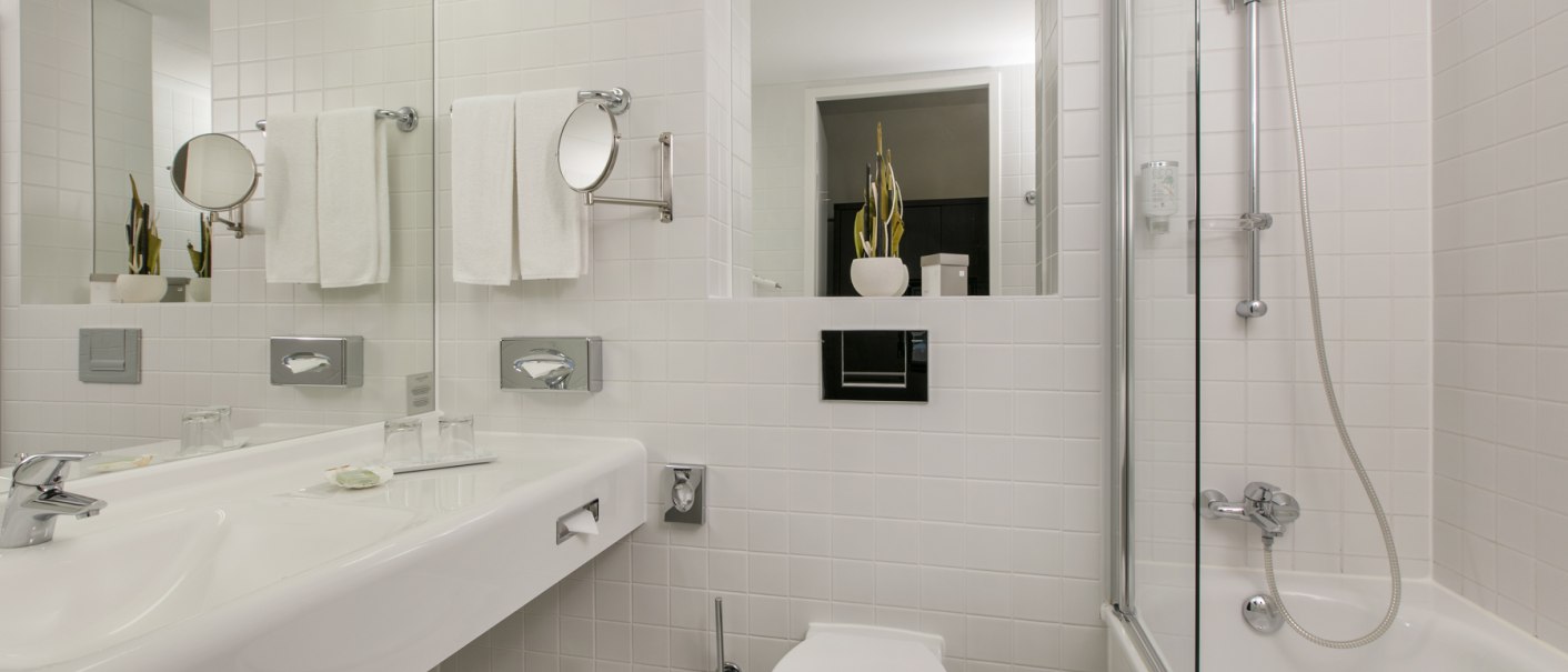 Modernes Badezimmer mit wei&szlig;en Fliesen, Waschbecken, Toilette, Badewanne und Spiegel. Handt&uuml;cher und Pflanzen als Dekoration., &copy; ARCOTEL Hotels