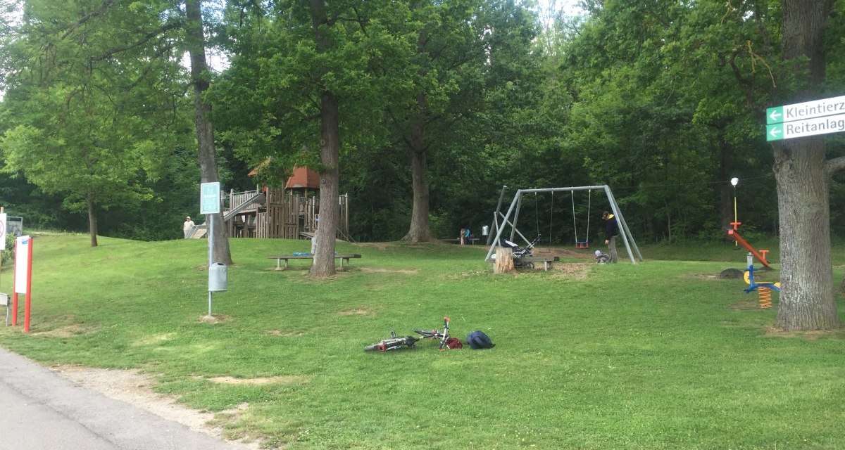 Spielplatz mit Schaukeln und Klettergerüst, umgeben von Bäumen. Ein Fahrrad liegt auf dem Rasen. Wegweiser zu Kleintierzoo und Reitanlage., © www.pro-cycl.de Spielplatz mit Schaukeln und Klettergerüst, umgeben von Bäumen. Ein Fahrrad liegt auf dem Rasen. Wegweiser zu Kleintierzoo und Reitanlage., © www.pro-cycl.de