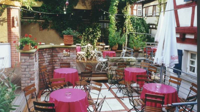 Ein gemütlicher Café-Innenhof mit Holztischen und -stühlen, rosa Tischdecken, umgeben von Pflanzen und Fachwerkhäusern., © 's Café Ein gemütlicher Café-Innenhof mit Holztischen und -stühlen, rosa Tischdecken, umgeben von Pflanzen und Fachwerkhäusern., © 's Café