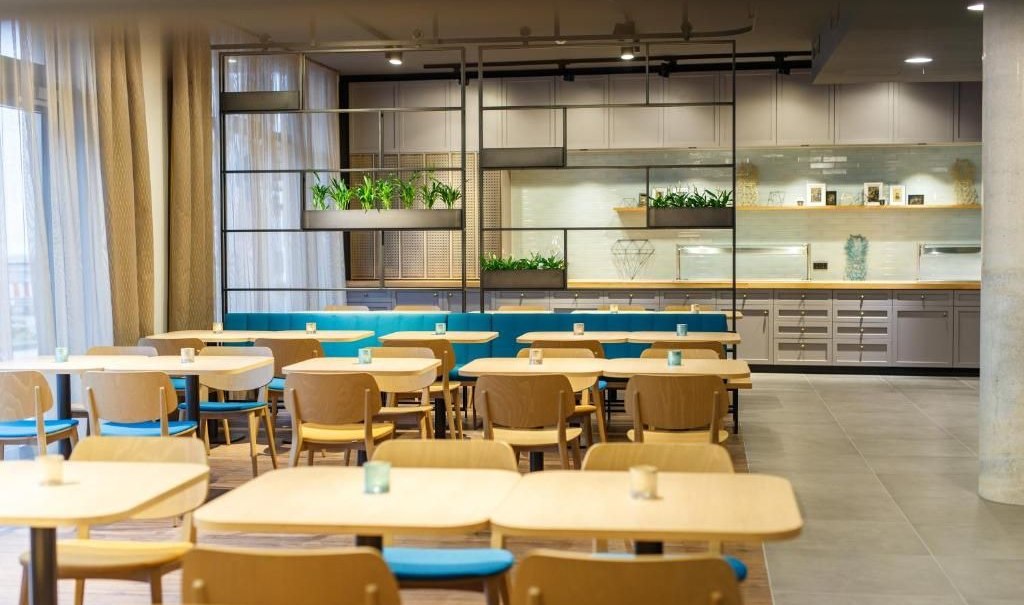 Moderner Speisesaal mit Holztischen und blauen Stühlen. Im Hintergrund sind Pflanzen und Regale zu sehen. Helle, einladende Atmosphäre., © Arthotel ANA Momentum