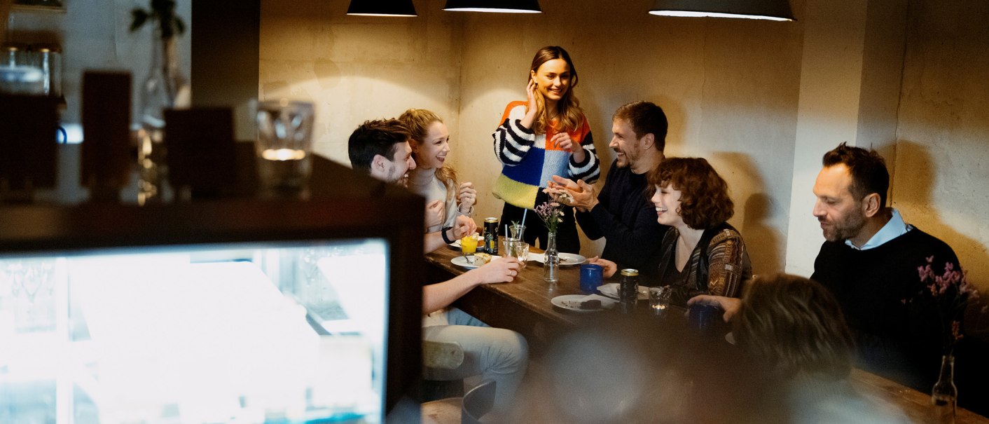 Eine Gruppe von Menschen sitzt in einem Café um einen Tisch und lacht. Eine Frau steht und spricht mit den anderen. Die Atmosphäre ist gemütlich., © Stuttgart Marketing GmbH, Alwin Maigler