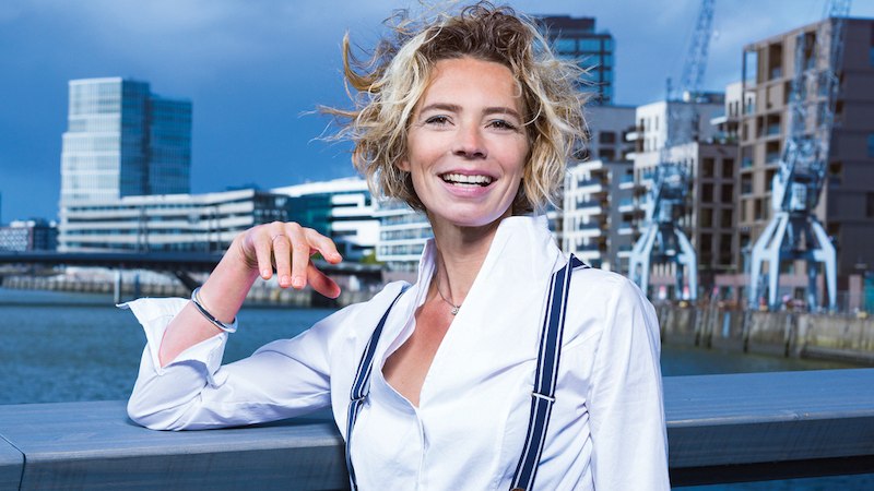 Frau mit lockigem Haar und wei&szlig;em Hemd l&auml;chelt vor moderner Stadtlandschaft mit Wasser und Geb&auml;uden., &copy; Kultur- und Sportamt Stadt Bietigheim-Bissingen