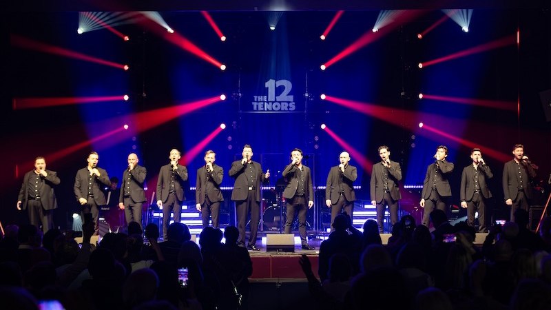 Zwölf Tenöre stehen auf einer Bühne, singen in Mikrofone. Bunte Lichteffekte und ein Publikum im Vordergrund. Schriftzug 'The 12 Tenors' im Hintergrund., © die Halle Kulturinitiative e.V.