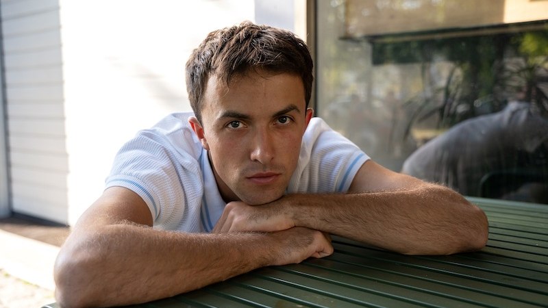 Ein Mann in einem weißen T-Shirt lehnt sich auf einen grünen Tisch und schaut direkt in die Kamera. Im Hintergrund sind unscharfe Pflanzen zu sehen., © Kulturhaus Schwanen