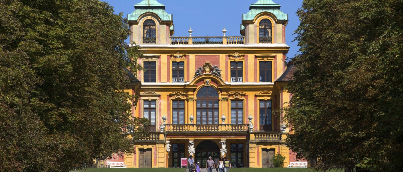 Barockes Schloss mit gelber Fassade und grünen Dächern, umgeben von Bäumen. Menschen stehen vor dem Eingang., © Tourismus & Events Ludwigsburg Barockes Schloss mit gelber Fassade und grünen Dächern, umgeben von Bäumen. Menschen stehen vor dem Eingang., © Tourismus & Events Ludwigsburg