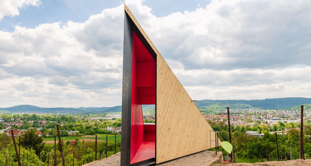 Moderne Holzstruktur mit rotem Innenraum auf einem Hügel. Im Hintergrund eine weite Landschaft mit Hügeln und Wolken am Himmel., © Authentic Studios Moderne Holzstruktur mit rotem Innenraum auf einem Hügel. Im Hintergrund eine weite Landschaft mit Hügeln und Wolken am Himmel., © Authentic Studios