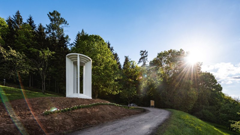 Weiße Säulenstruktur auf einem Hügel neben einem Weg, umgeben von Bäumen und Sonnenschein., © Authentic Studios