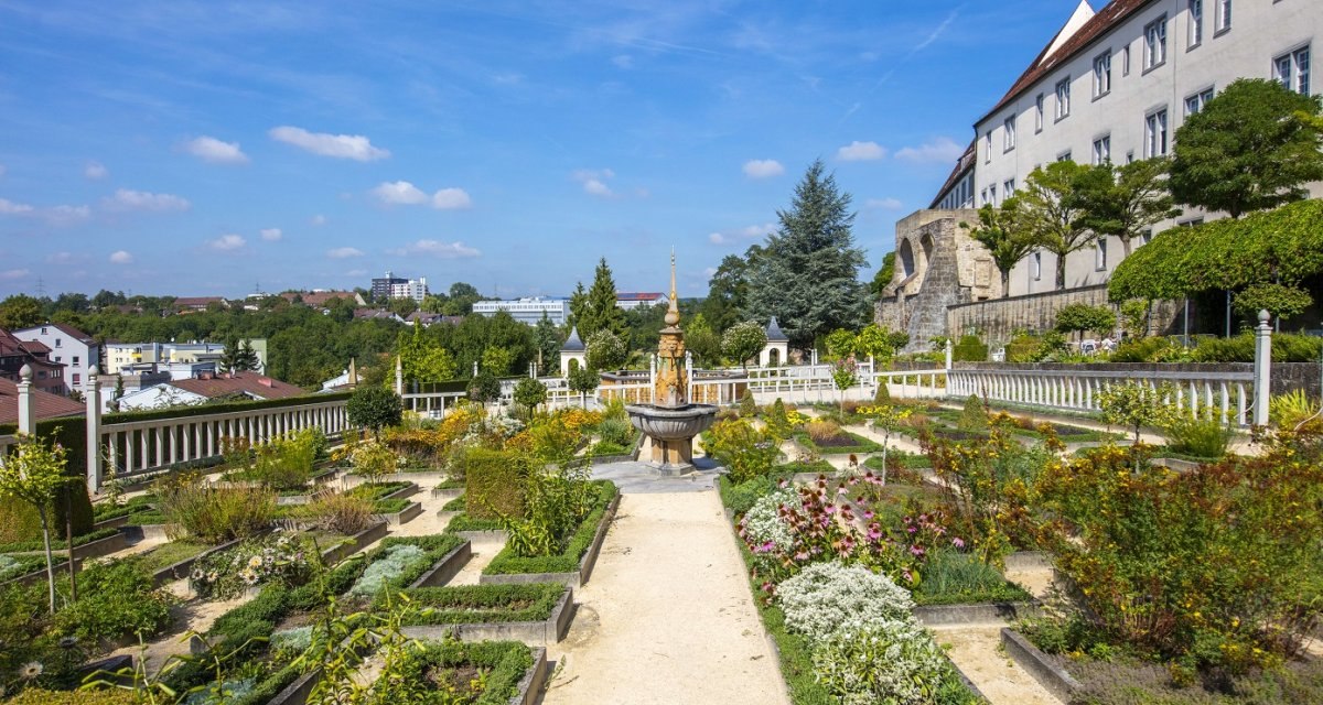 Ein gepflegter Garten mit symmetrischen Blumenbeeten und einem zentralen Brunnen, umgeben von historischen Geb&auml;uden und B&auml;umen unter blauem Himmel., &copy; Stuttgart Marketing GmbH Achim Mende