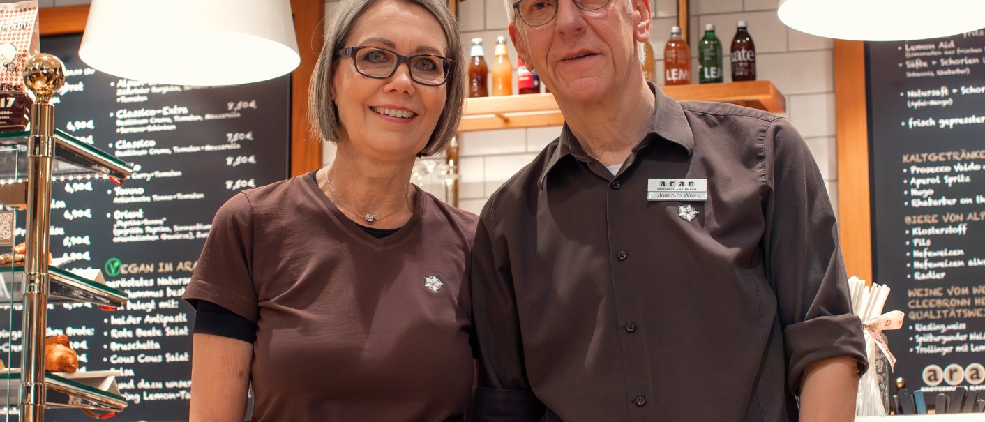 Zwei Personen stehen lächelnd vor einem Café namens 'aran', das Brotgenuss und Kaffeekultur anbietet., © Cafe aran Zwei Personen stehen lächelnd vor einem Café namens 'aran', das Brotgenuss und Kaffeekultur anbietet., © Cafe aran