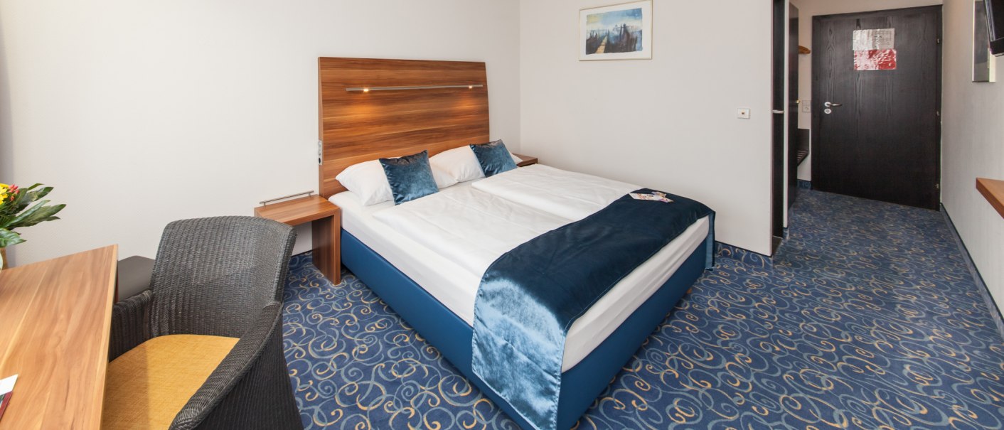 Ein Hotelzimmer mit Doppelbett, blauem Teppich und Holzmöbeln. Ein Schreibtisch mit Stuhl und ein Bild an der Wand sind sichtbar., © Novum Management GmbH