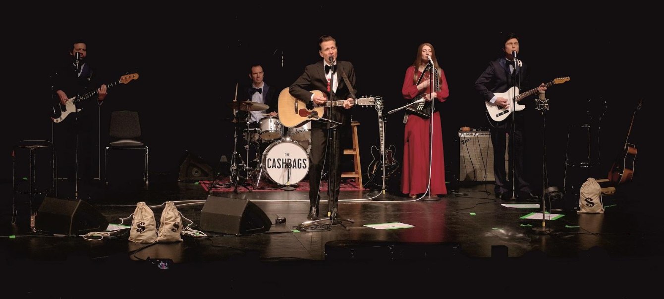 Die Band The Cashbags performt auf der Bühne. Fünf Musiker spielen Gitarre, Schlagzeug und singen. Im Vordergrund sind Geldsäcke zu sehen., © © Roland März
