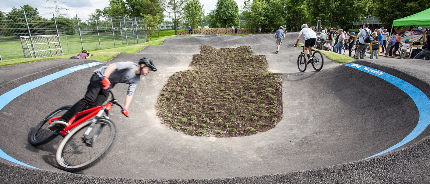 Fahrradfahrer auf einem Pumptrack in Sindelfingen, umgeben von Zuschauern und Bäumen. Die Strecke ist asphaltiert und wellig., © Fotoknobi