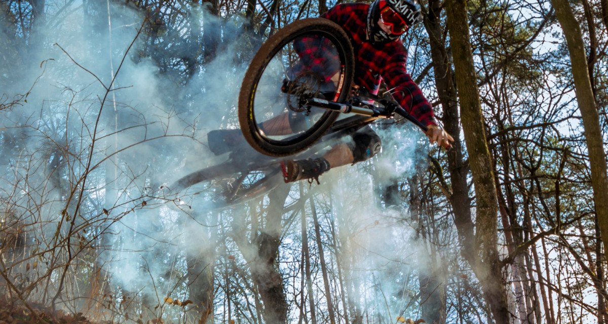 Ein Mountainbiker in rotem Karohemd springt durch Rauch im Wald. Die Szene ist dynamisch und zeigt herbstliche Bäume im Hintergrund., © Natur.Nah. Schönbuch & Heckengäu
