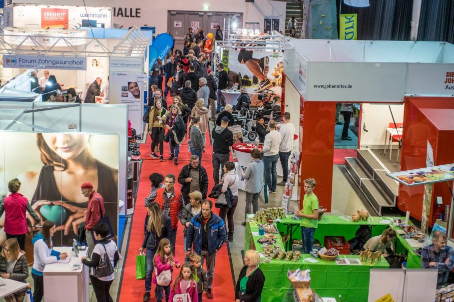 Menschen auf der vitawell Messe mit Ständen zu Zahngesundheit und anderen Themen. Roter Teppich führt durch die Halle.