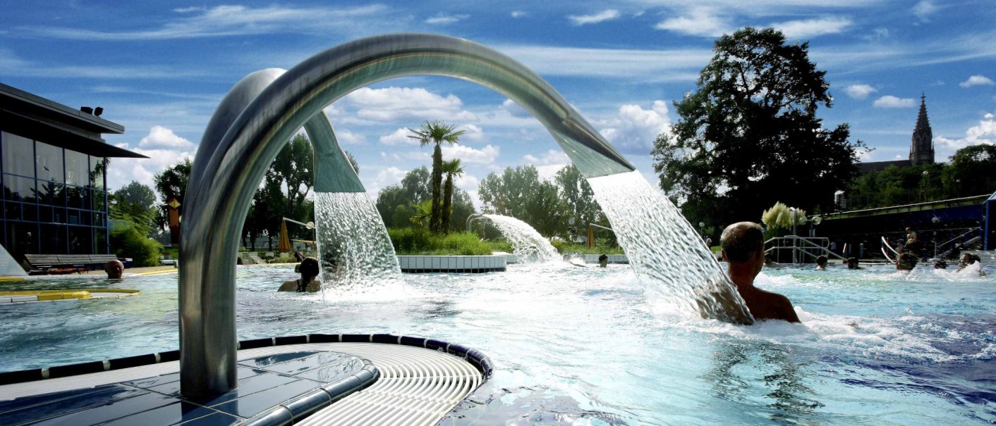 Ein Außenpool mit Wasserfontänen und Palmen, umgeben von Bäumen und einem Gebäude. Menschen genießen das Wasser unter blauem Himmel., © Stuttgarter Bäder