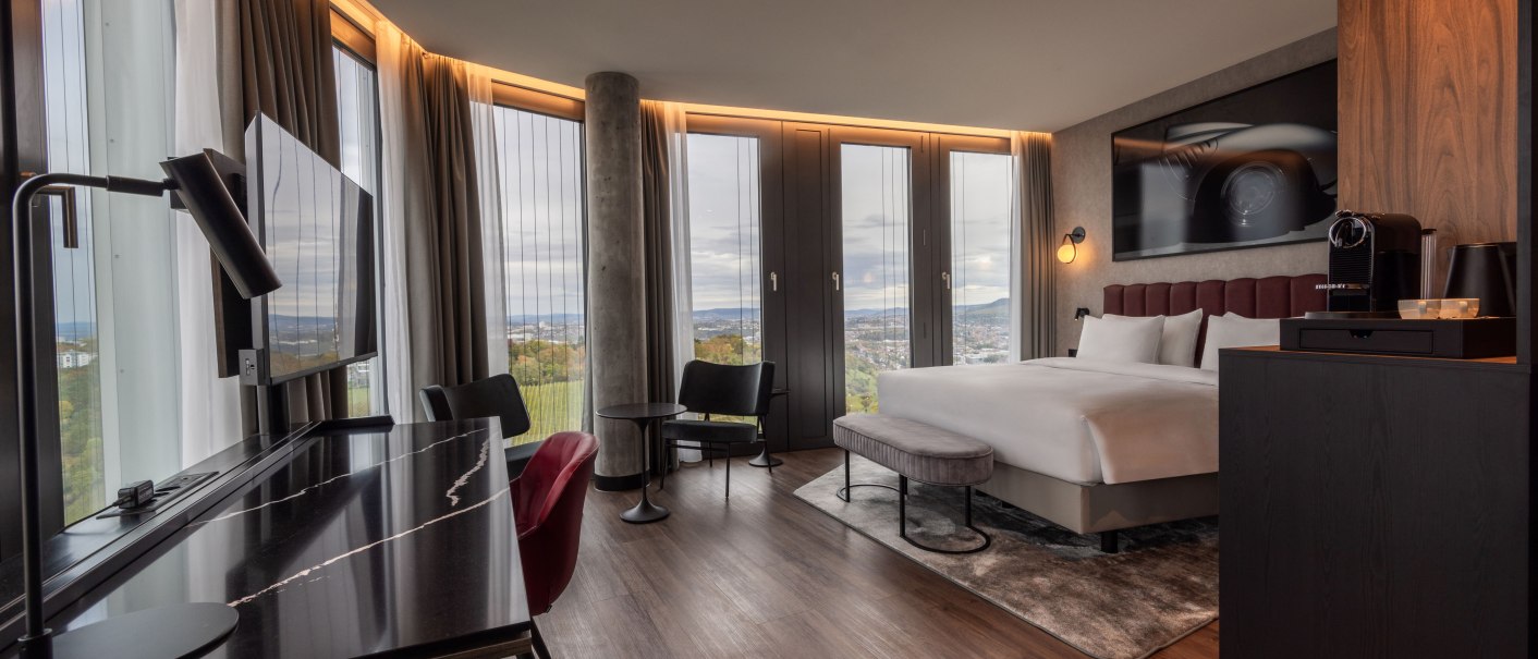 Modernes Eckzimmer mit Panoramafenstern, Doppelbett, Schreibtisch, Sessel und Kaffeemaschine. Elegantes Design mit Holzboden und stilvoller Beleuchtung., © Radisson Hotel Group