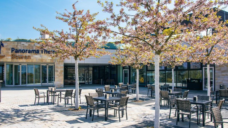 Eingangsbereich der Panorama Therme Beuren mit blühenden Bäumen und leeren Sitzplätzen im Freien. Die Sonne scheint an einem klaren Tag., © Stuttgart-Marketing GmbH, Sarah Schmid