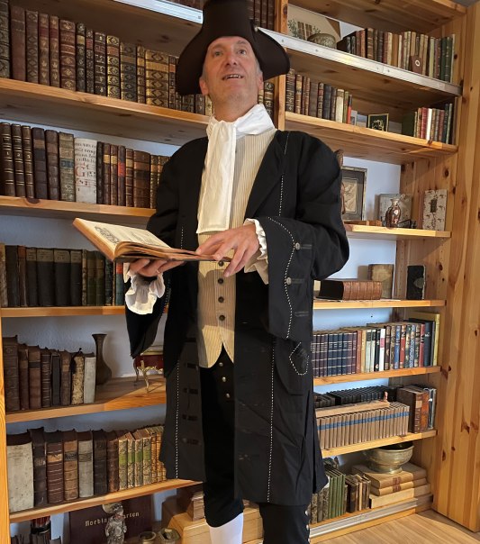 Eine Person in historischer Kleidung mit Dreispitz und R&uuml;schenhemd steht vor einem B&uuml;cherregal und h&auml;lt ein aufgeschlagenes Buch., &copy; Gerd Jenner