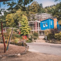 Spielplatz mit buntem Klettergerüst, Holzspänen und einem blauen Gebäude im Hintergrund, umgeben von Bäumen., © Stadt Welzheim