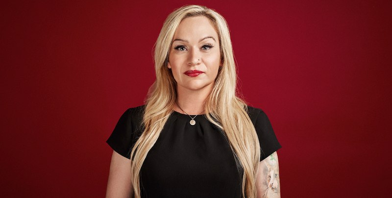 Eine Frau mit langen blonden Haaren und schwarzem Kleid steht vor einem roten Hintergrund. Sie hat ein Tattoo am Arm und trägt roten Lippenstift., © Scala Kultur Live gGmbH