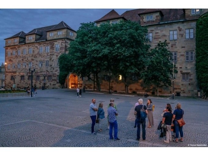 Menschen stehen auf einem gepflasterten Platz vor einem historischen Geb&auml;ude bei D&auml;mmerung. Im Hintergrund sind B&auml;ume und beleuchtete Fenster zu sehen., &copy; Stuttgart Marketing GmbH