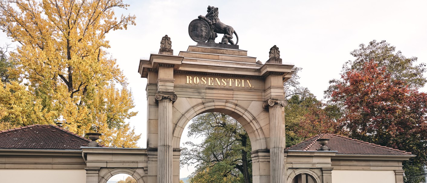 Historisches Tor mit 'Rosenstein'-Schriftzug, flankiert von Gebäuden. Herbstliche Bäume im Hintergrund, zwei Personen spazieren., © Stuttgart-Marketing GmbH, Christoph Düpper